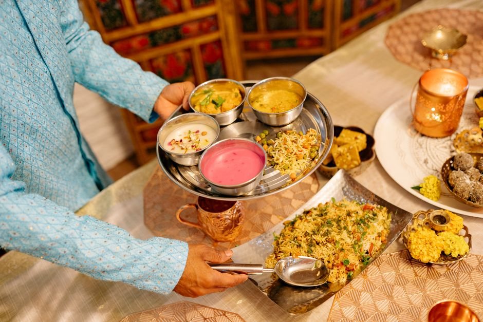 Server in a blue kurta carrying a steel thali of vegetarian rice, dal, curries and sweets at a festive Indian meal