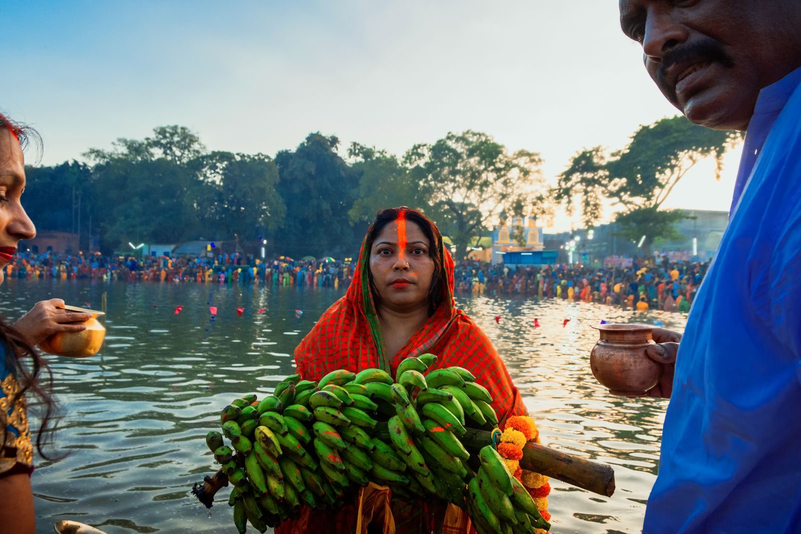 Woman in an orange sari with sindoor on her forehead holding a large bunch of green bananas with marigolds at a crowded Chhath ghat