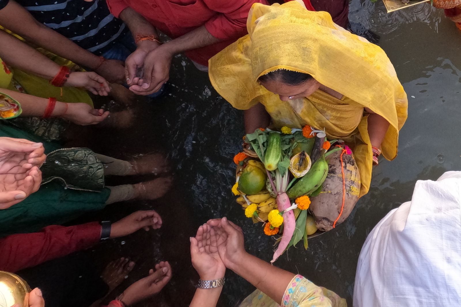 Woman in a yellow sari standing in a river holding a soop of radish, cucumber, bananas, gagal lemon, coconut and a diya while family members hold out cupped hands