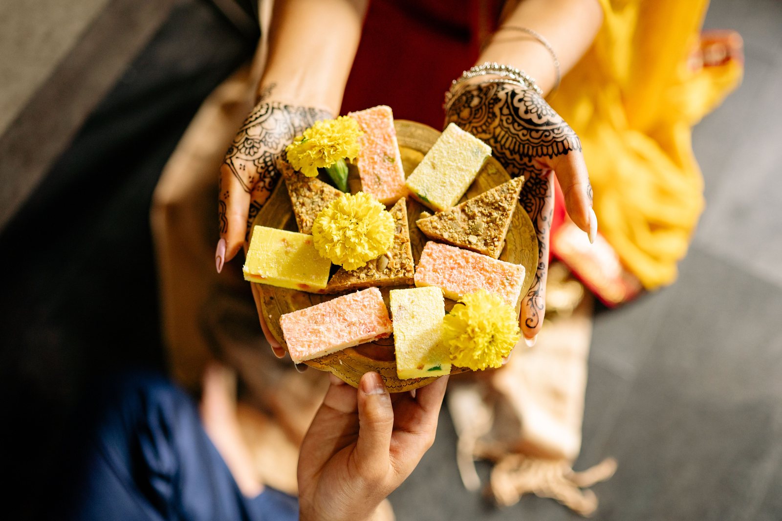 Hands with mehendi passing a brass plate of pink, yellow and green barfi decorated with yellow marigold flowers