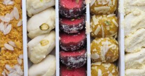 Open box of assorted Indian sweets in rows: almond-topped barfi, white milk rolls with pistachio, red coconut rolls and saffron ladoo