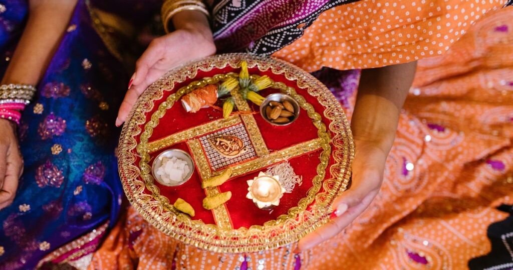 Woman in a bandhani saree holding a red and gold decorated thali with almonds, mishri, a lit diya and kalava thread